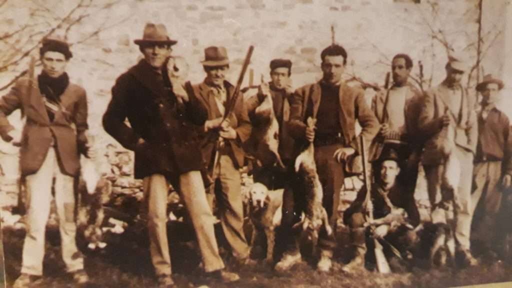 Foto storica color seppia: un gruppo di uomini di Urzano in posa davanti a un muro di pietra